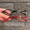 Une main tient des lunettes de mode ovales rouges rétro sur un fond de brique grise.