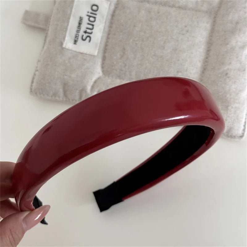 A close-up of a glossy maroon PU leather fashion headband being held against a light gray fabric background.