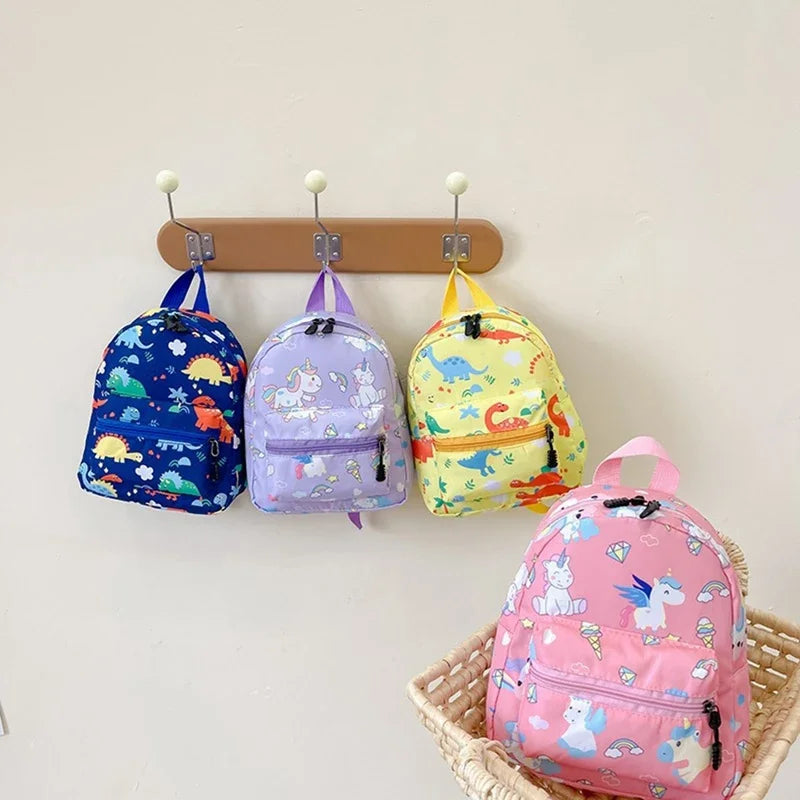 Three colorful kids backpacks featuring dinosaur and unicorn patterns hanging on a wall