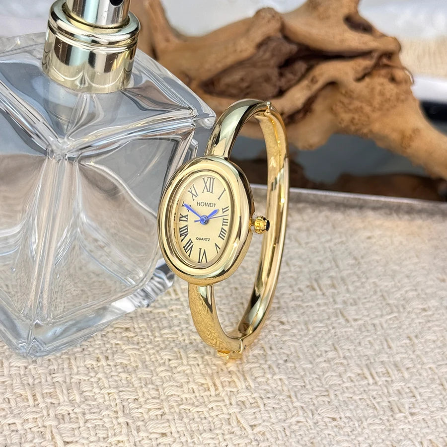 A gold-toned quartz watch with a clear face and Roman numerals is displayed next to a glass perfume bottle.