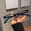 A hand holds a pair of classic black square eyeglasses with clear lenses against a light background.