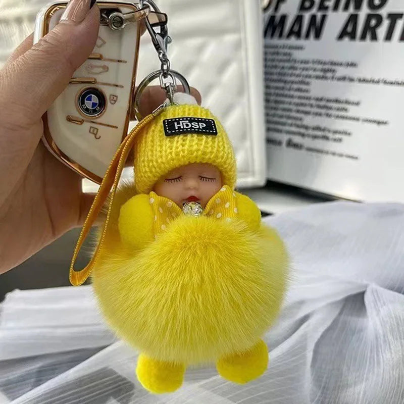 A yellow fluffy cartoon plush doll keychain with a silver charm is held in a hand.