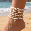 A wrist wearing a stack of white and gold beaded bracelets, set against a beach background.