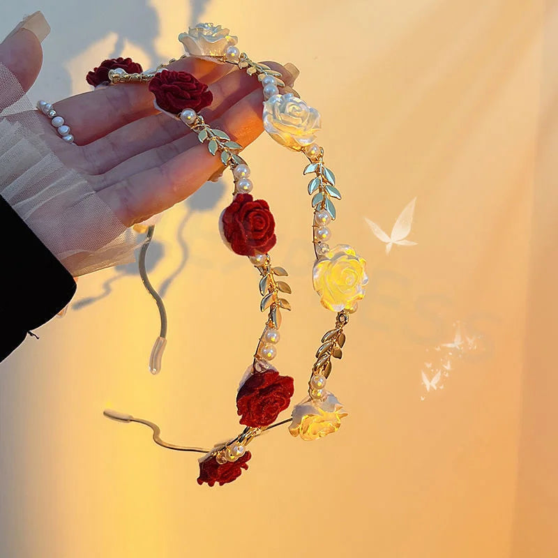 A hand holds an elegant rose vine headband hair accessory featuring red and yellow roses and pearls against a soft sunset bac