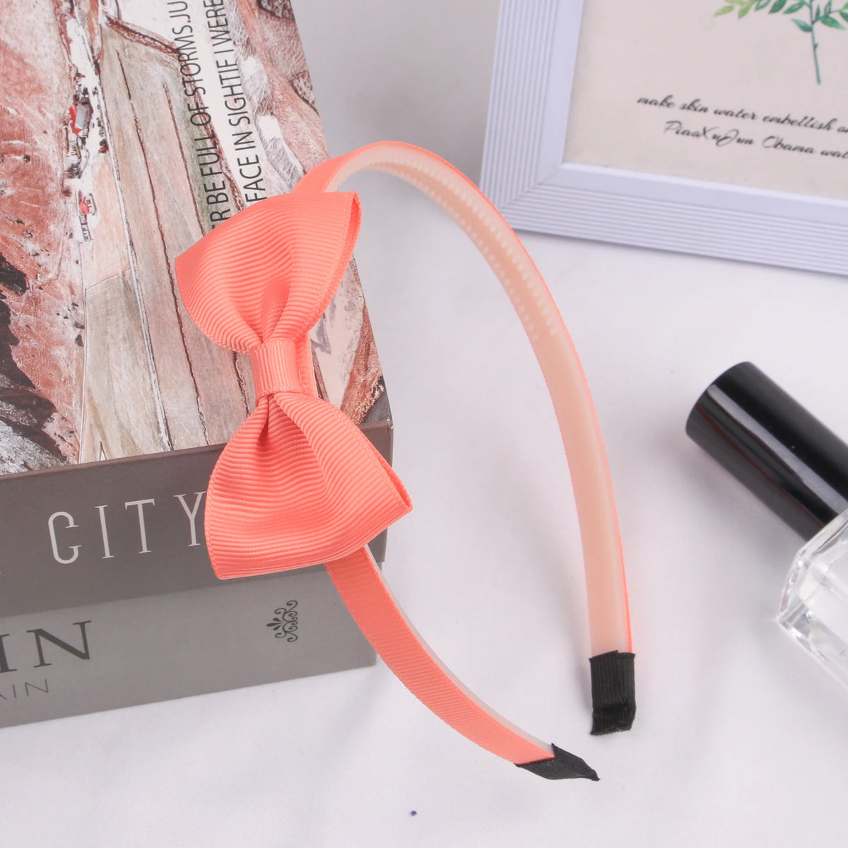 A pink grosgrain ribbon hair bow is displayed next to a book and a cosmetic bottle.