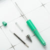 A green beadable ballpoint pen and a silver spring are shown on a white surface.