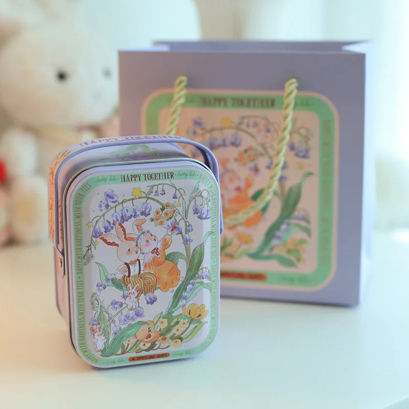 A decorative tinplate candy box with a carrying loop featuring floral designs and a light purple background.