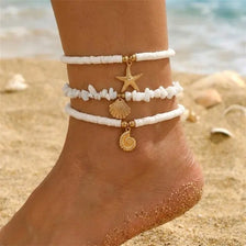 Un conjunto de tobilleras de cuentas de arroz blancas con adornos dorados usadas en el tobillo de una persona en una playa de