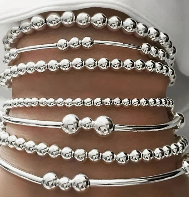 A collection of silver minimalist round beaded bracelets stacked on a person's wrist.