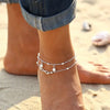 A close-up of a boho silver-plated friendship anklet worn on a woman's ankle against a sandy background.