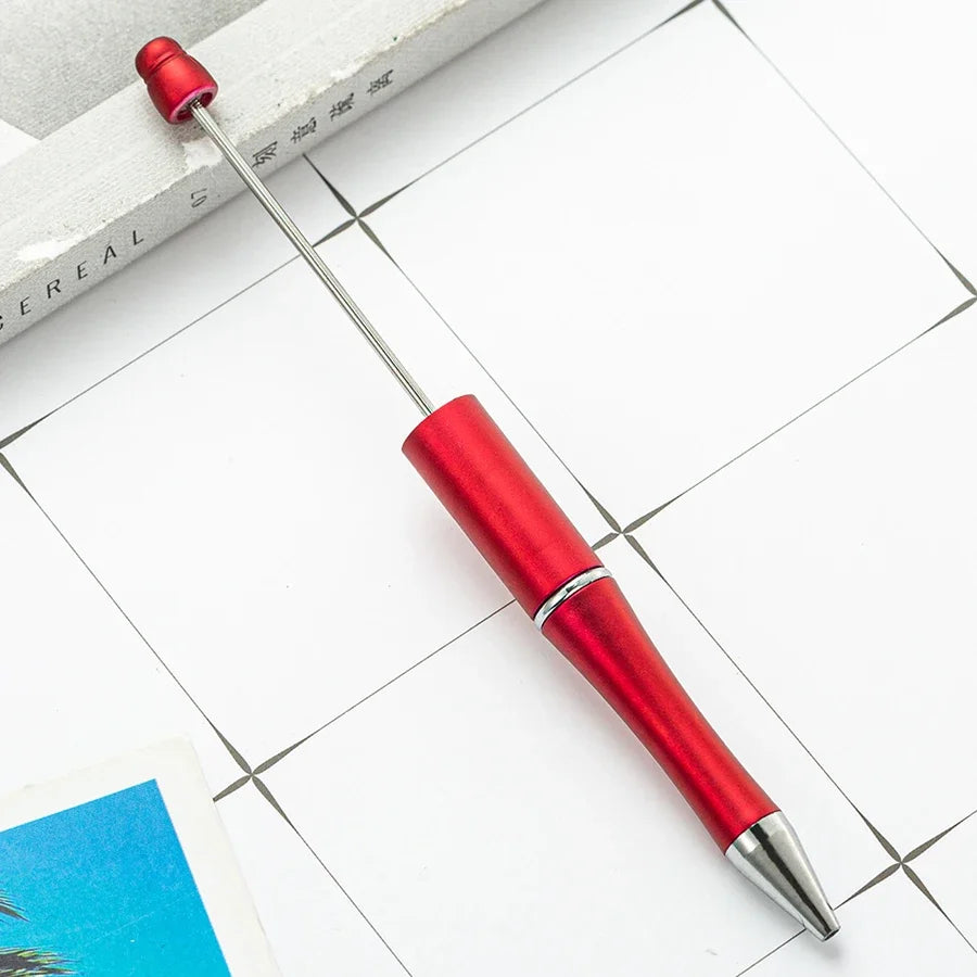 A red beadable ballpoint pen is shown lying on a white surface next to a book.
