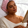A woman wearing a pink satin hair cap is reading a book in a bright, indoor setting.