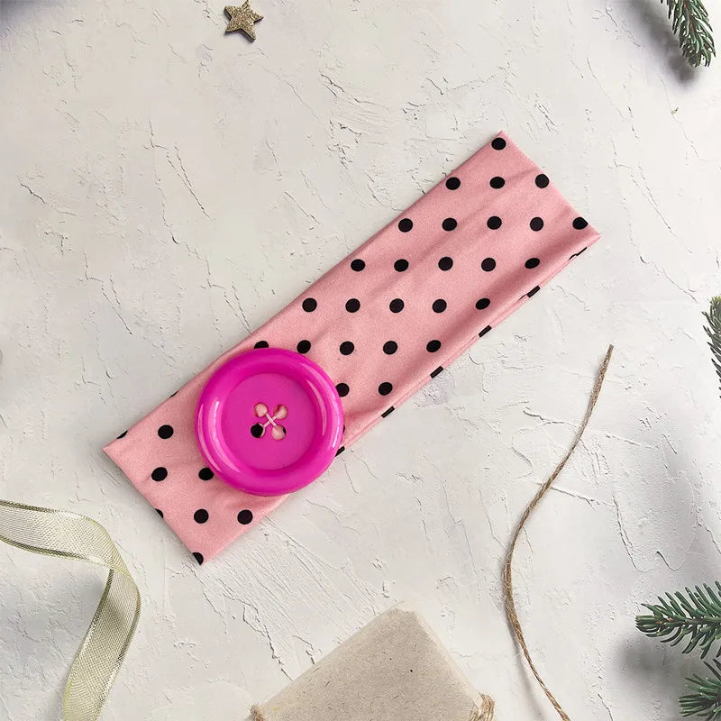 A pink polka dot button headband lying on a white surface with a gold ribbon and pine greenery.
