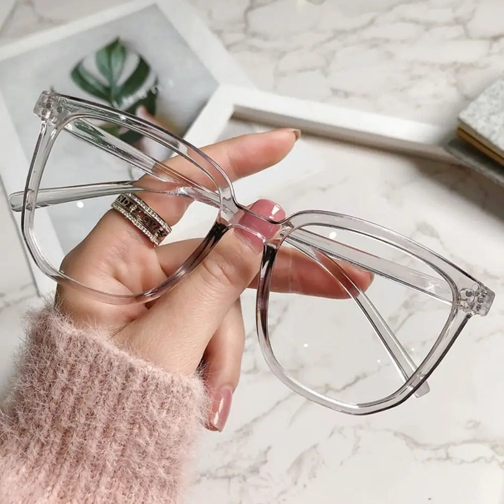 A hand holds a pair of clear, large square anti-blue light spectacles against a marble background.