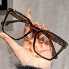 A hand holds a pair of elegant geometric reading glasses with dark brown frames and transparent lenses.