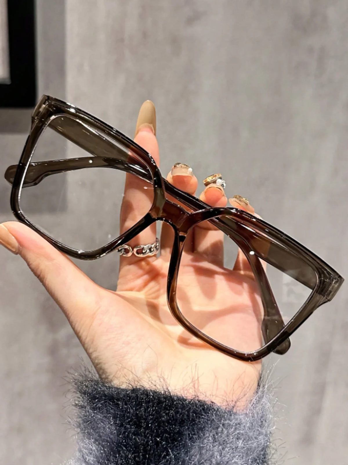 A hand holds a pair of elegant geometric reading glasses with dark brown frames and transparent lenses.