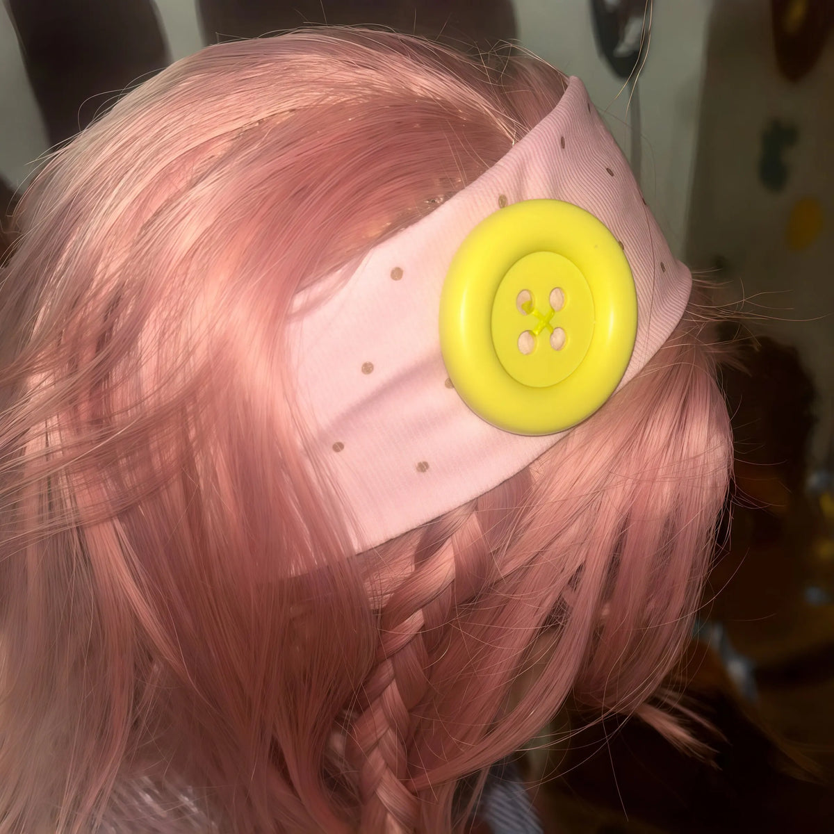 A pink hair styled in braids wears a light pink headband with a yellow polka dot button detail.