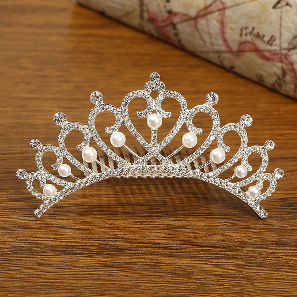 A sparkling crystal princess tiara hair comb adorned with pearls resting on a wooden surface.