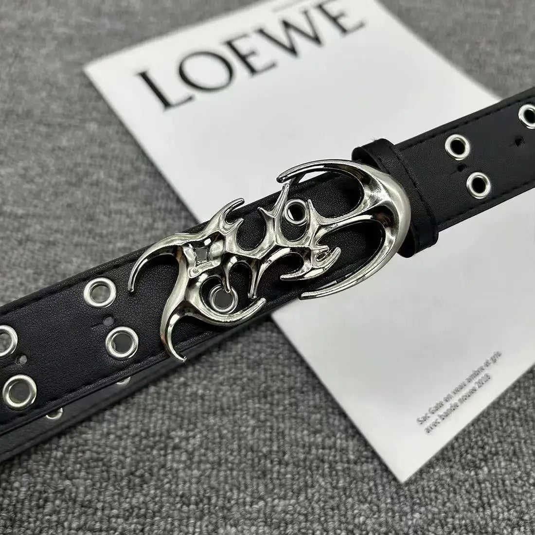 A black leather belt with a silver skull buckle is displayed on a white card.
