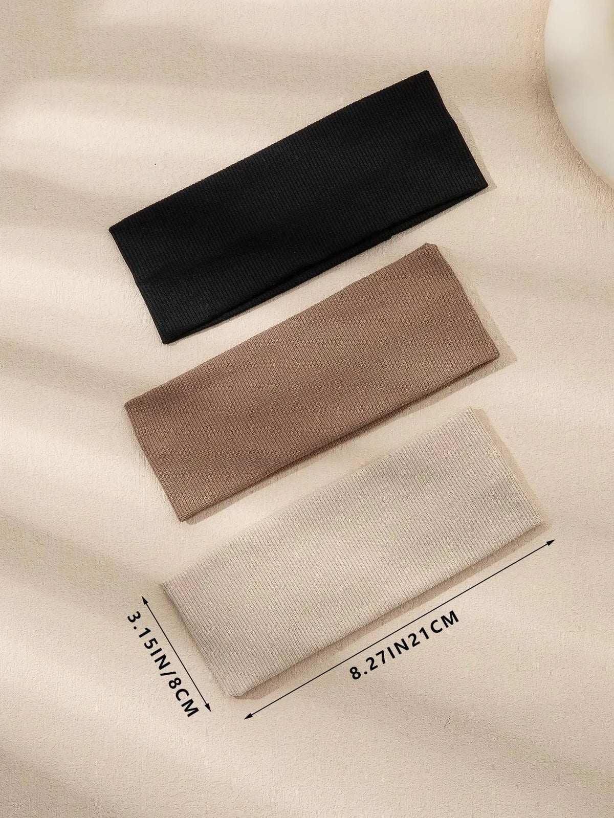 Three solid color fashion headbands in black, beige, and cream laid diagonally on a light background.