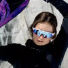 A young person wearing blue mirrored cycling sunglasses with a white frame against a light background.