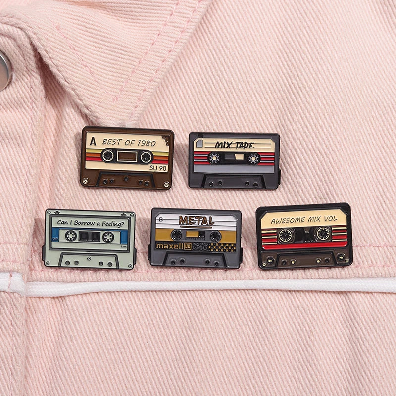 A collection of four retro cassette enamel pins featuring different tape designs and labels on a pink fabric background.