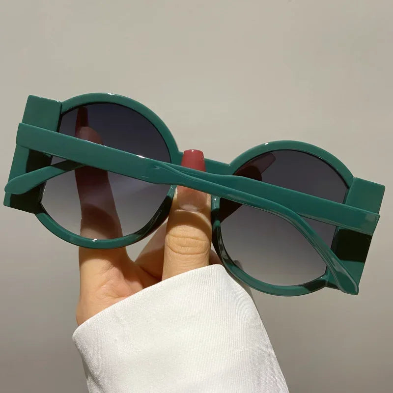 A hand holds a pair of round double color fashion sunglasses with green frames and dark lenses.