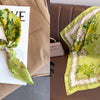 A light green floral print silk scarf tied in a bow displayed on a white card against a tan blazer.