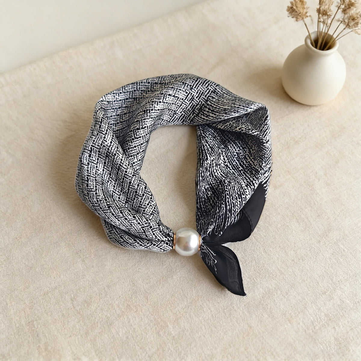 A black and white plaid silk scarf tied with a satin bow and a pearl accent, displayed on a light beige surface.