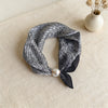 A black and white plaid silk scarf tied with a satin bow and a pearl accent, displayed on a light beige surface.