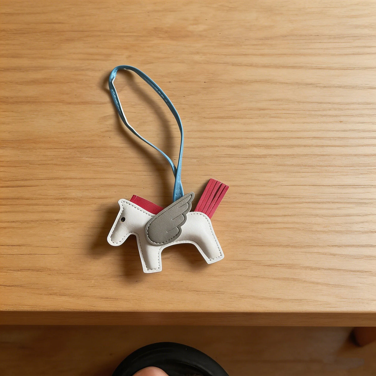 A white horse pendant charm with gray wings and a blue cord resting on a wooden surface.