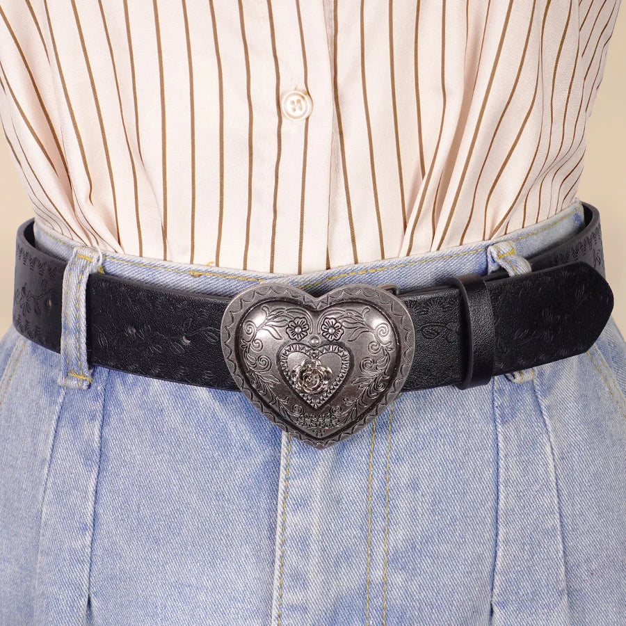 A close-up of a heart-shaped retro embossed black leather belt worn over light pink striped shirt and blue jeans.