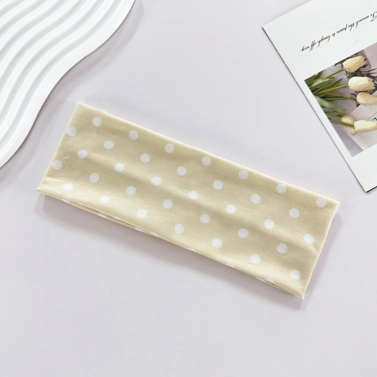 A beige fabric headband with white polka dots is displayed on a light surface next to a card.