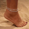 A close-up of a person's ankle showing layered gold-plated twisted bracelets worn around the ankle.