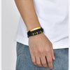 Nahaufnahme von einem Handgelenk mit schwarzem und goldenem geflochtenem Lederarmband vor weißem Hemd und blauen Jeans.