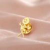 A gold-colored punk stickman brooch with a smiling face and outstretched arms resting on pink fabric.