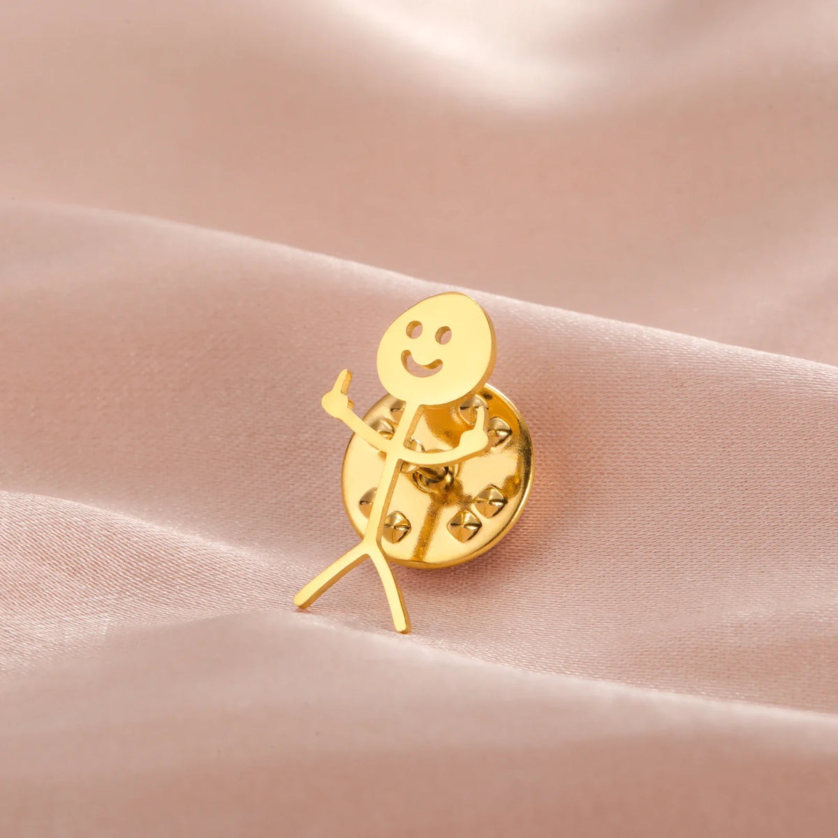 A gold-colored punk stickman brooch with a smiling face and outstretched arms resting on pink fabric.