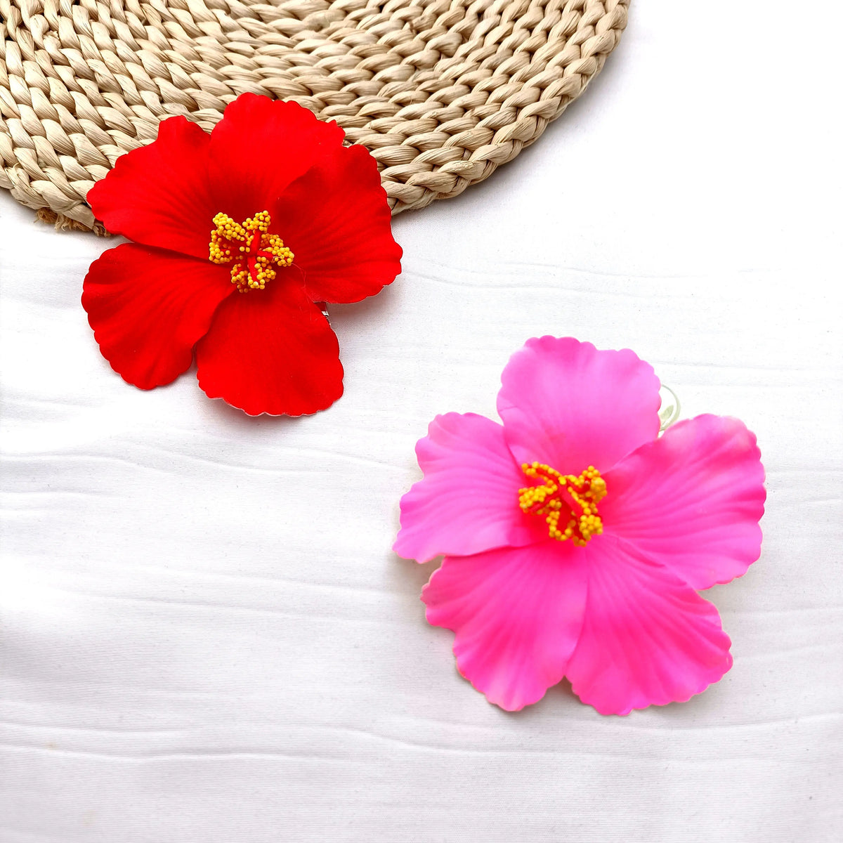 A vibrant red hibiscus flower rests on a woven mat next to a pink hibiscus flower on white fabric.