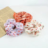 A set of three colorful satin fabric scrunchie headbands in shades of peach, pink, and cream are displayed next to a piece of