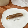 A light brown rubber braided ponytail holder is displayed on a white plate, with a measurement line indicating its length of 