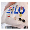 A light pink fabric features various decorative pins and badges, including a large blue text design and fruit motifs.