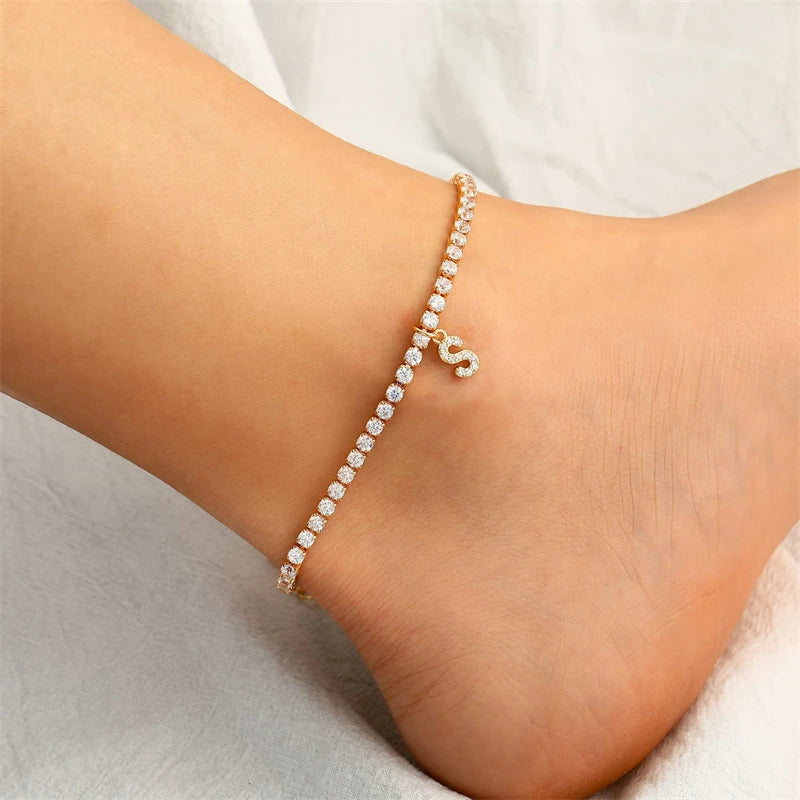 A gold-toned letter anklet bracelet with small clear stones worn on a person's ankle.