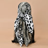 A black and white leopard print square satin scarf tied in a bow, displayed against a beige background.