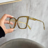 A hand holds a pair of elegant geometric reading glasses with transparent yellow-tinted lenses against a light gray backgroun