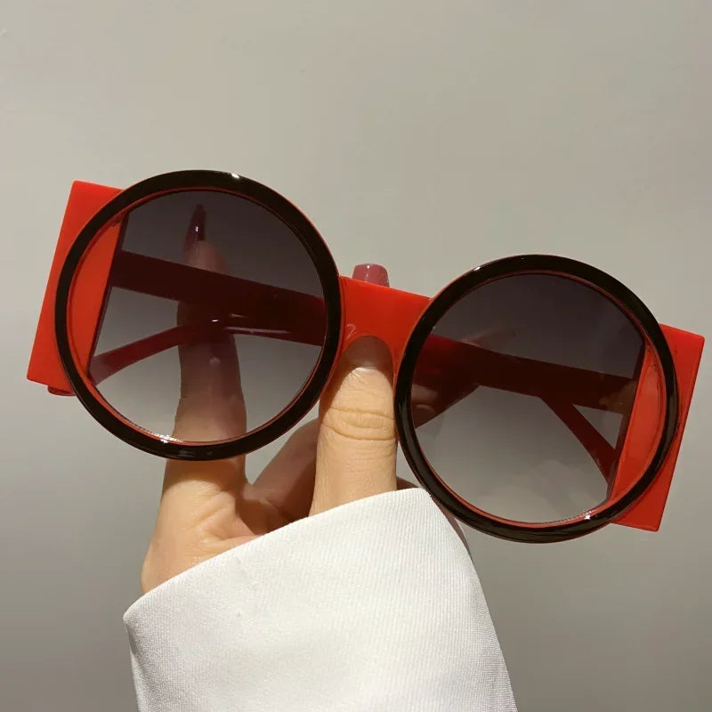 A hand holds a pair of round double color fashion sunglasses with red frames and dark lenses.