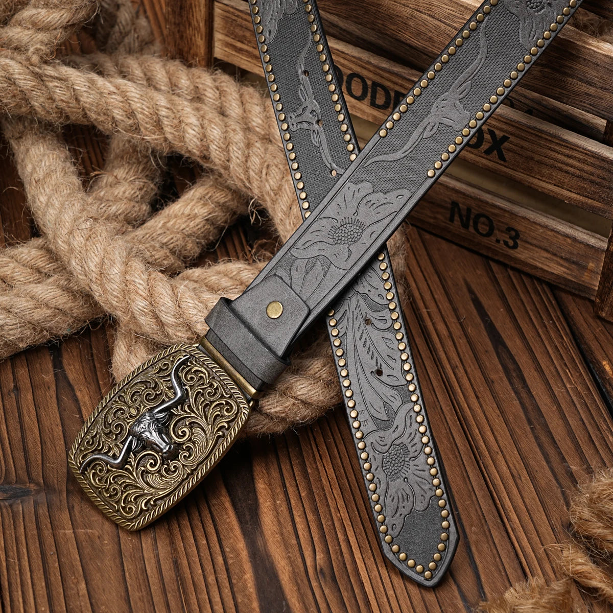 A dark leather belt with a floral engraving is laid across a thick rope on a wooden surface.