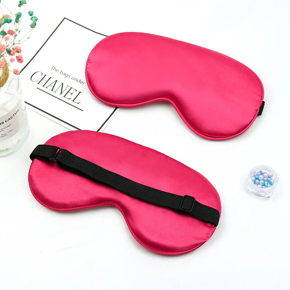 A vibrant pink silk sleep mask with a black strap is displayed next to a piece of paper.