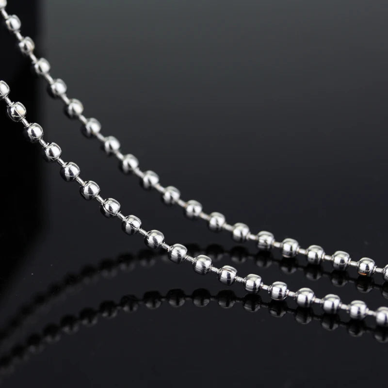 A close-up of a stainless steel pendant necklace with small silver beads, reflecting on a dark surface.