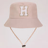 A light beige sun hat with a gold letter H embroidered in white faux fur on the front.