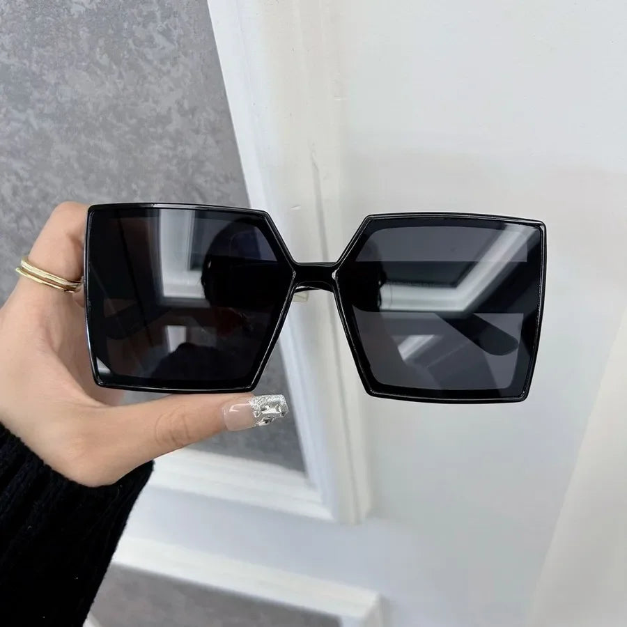 A pair of black mirror square sunglasses held by a hand against a white door frame.